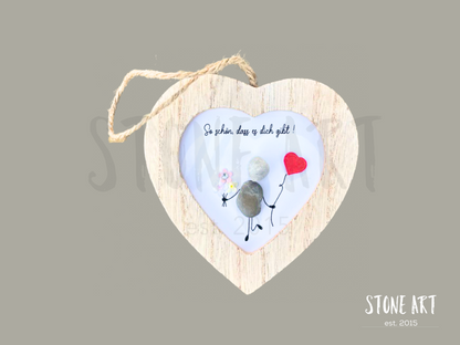  Steinbild mit der Aufschrift „So schön, dass es dich gibt“ in einem herzförmigen Holzrahmen. Zwei sitzende Figuren aus Kieselsteinen, ergänzt durch ein rotes Herz. Handgefertigte Wanddekoration zum Aufhängen.