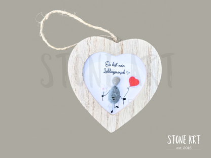 Steinbild mit der Aufschrift „Du bist mein Lieblingsmensch“ in einem herzförmigen Holzrahmen. Zwei Figuren aus Kieselsteinen sitzen nebeneinander, ergänzt durch ein kleines rotes Herz. Handgefertigte Wanddekoration zum Aufhängen.