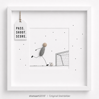 Steinbild mit einem Fußballspieler aus Kieselsteinen, der einen Ball auf ein gezeichnetes Tor schießt, Schriftzug Pass Shoot Score