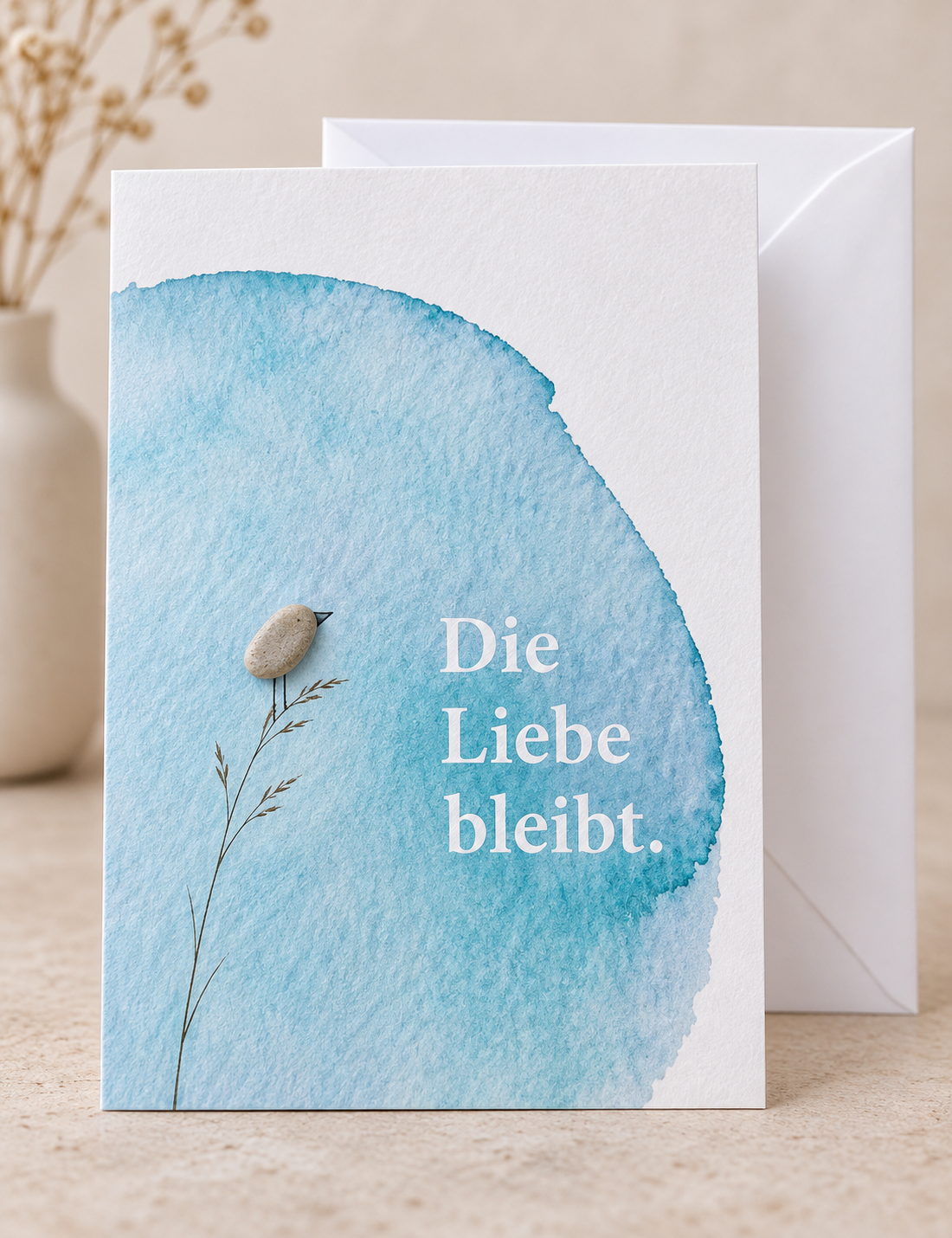 Handgemachte weiße Klappkarte mit Kieselstein-Vogel auf einem Grashalm vor blauem Aquarell, Spruch „Die Liebe bleibt.“ – Trauerkarte zur Anteilnahme, Grußkarte mit Umschlag